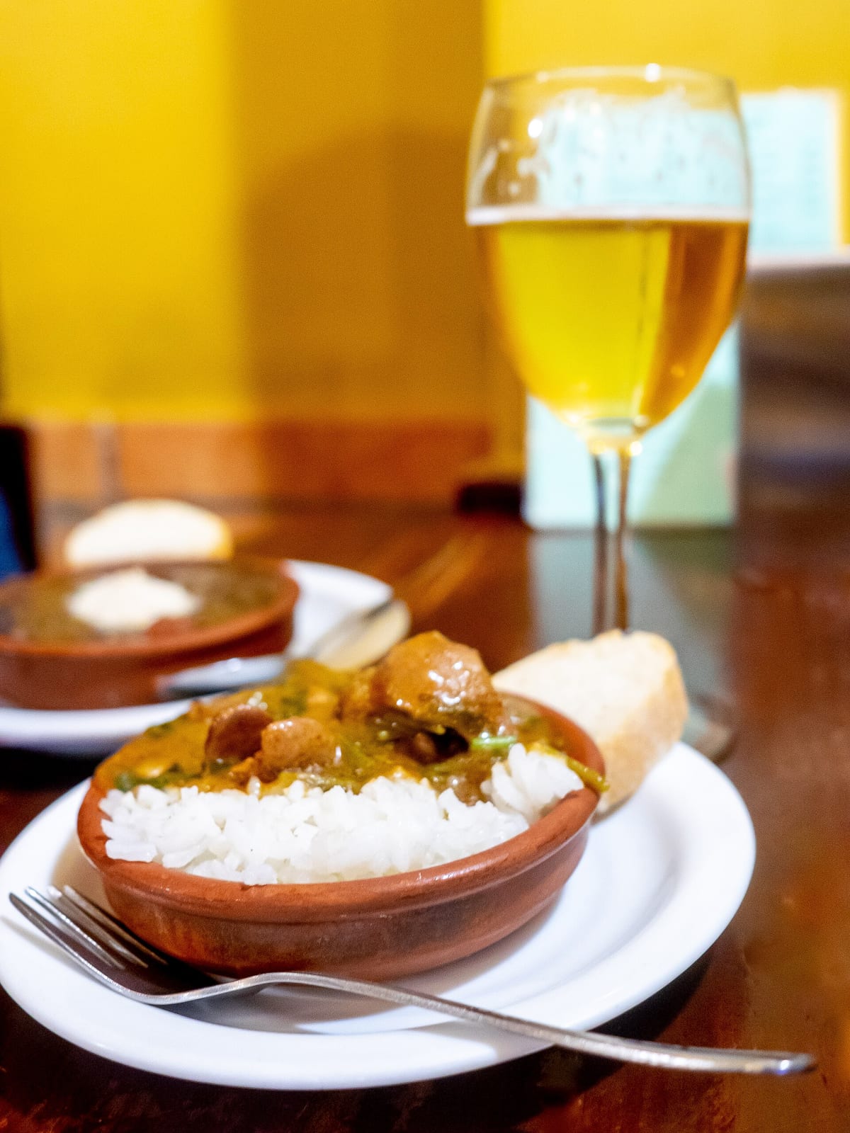 A small clay dish of meat stew over white rice with a piece of bread, beside a stemmed glass of beer on a wooden bar table in Granada