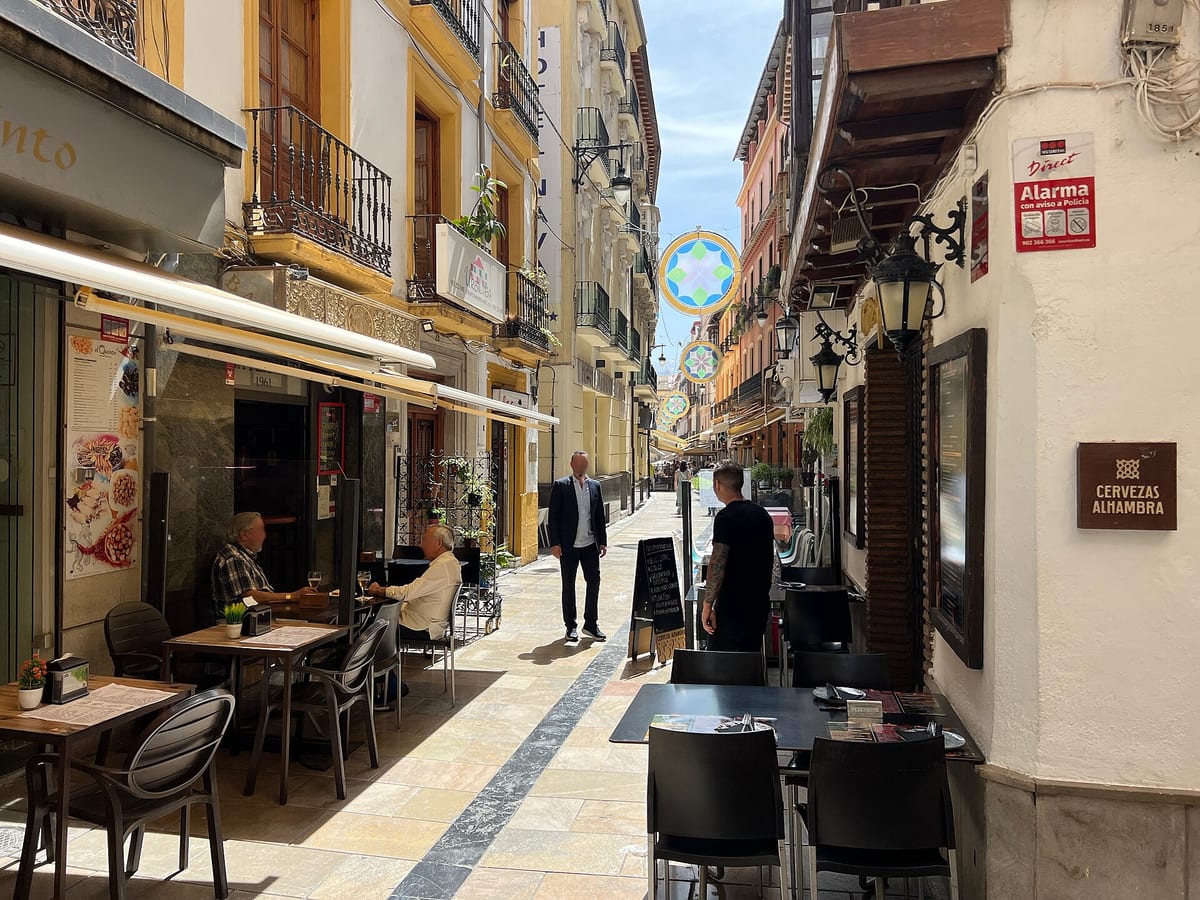 Calle Navas in central Granada on a sunny day: a narrow pedestrian street of yellow and white balconied buildings, lined with bar terraces of dark tables and chairs, a Cervezas Alhambra sign on the right