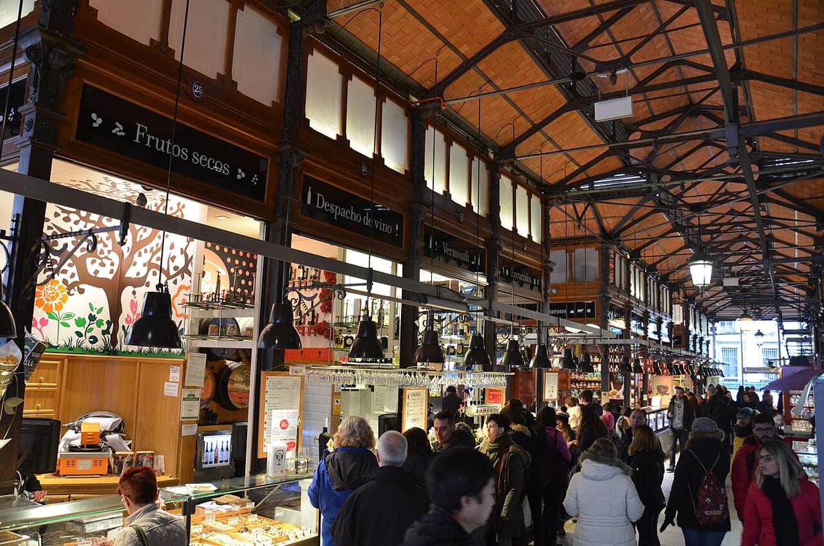 Shoppers along the wood-fronted stalls of the Mercado de San Miguel in Madrid, under a vaulted brick and iron roof