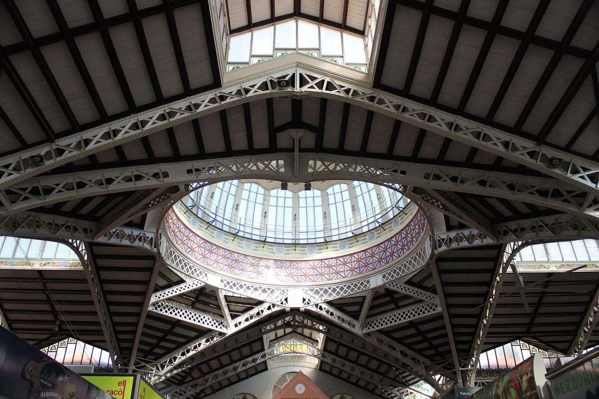Inside Valencia's Mercado Central, looking up at the iron roof trusses and the glass dome ringed with red and blue tilework, with market stall signs along the bottom of the frame