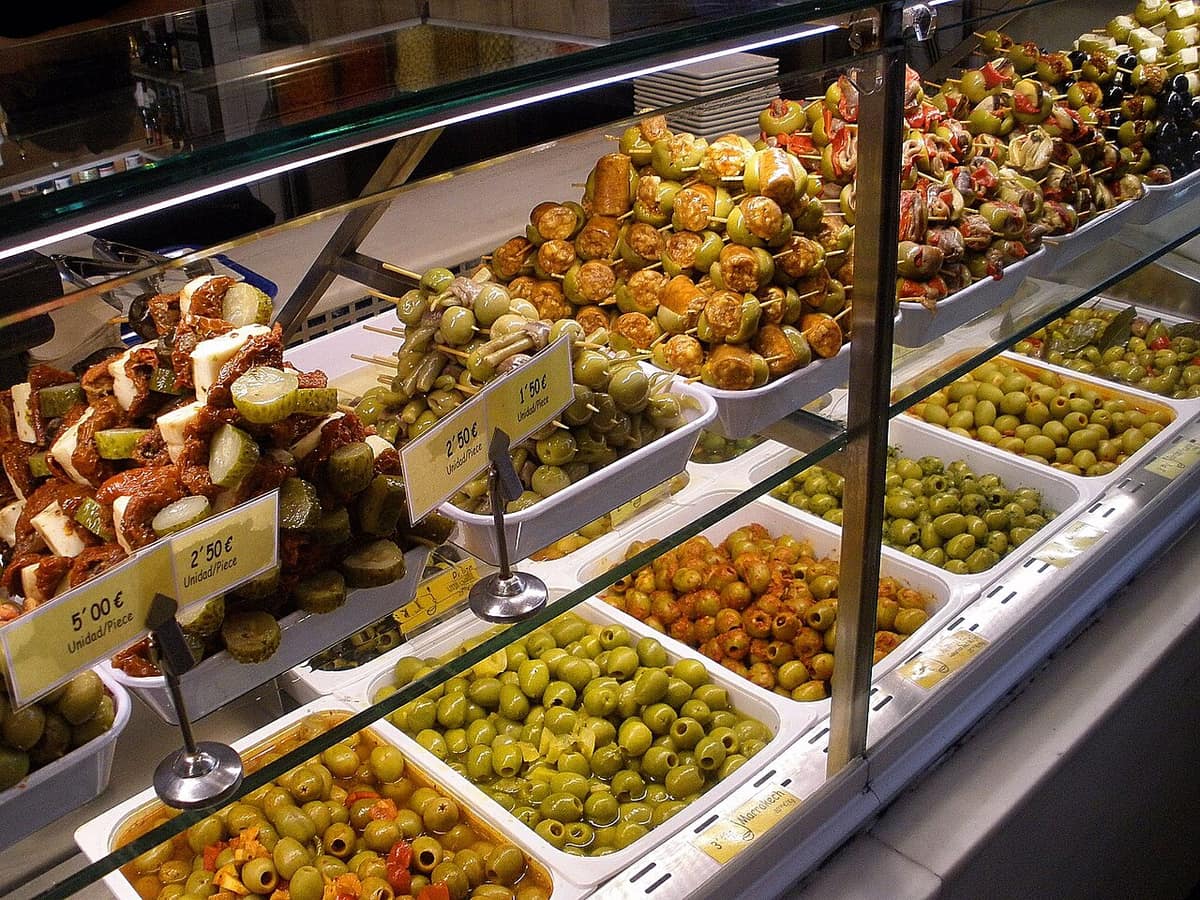 A glass counter at a Madrid market stall lined with trays of green olives and skewered pintxos of olives, pickles, peppers and anchovies, each priced per piece