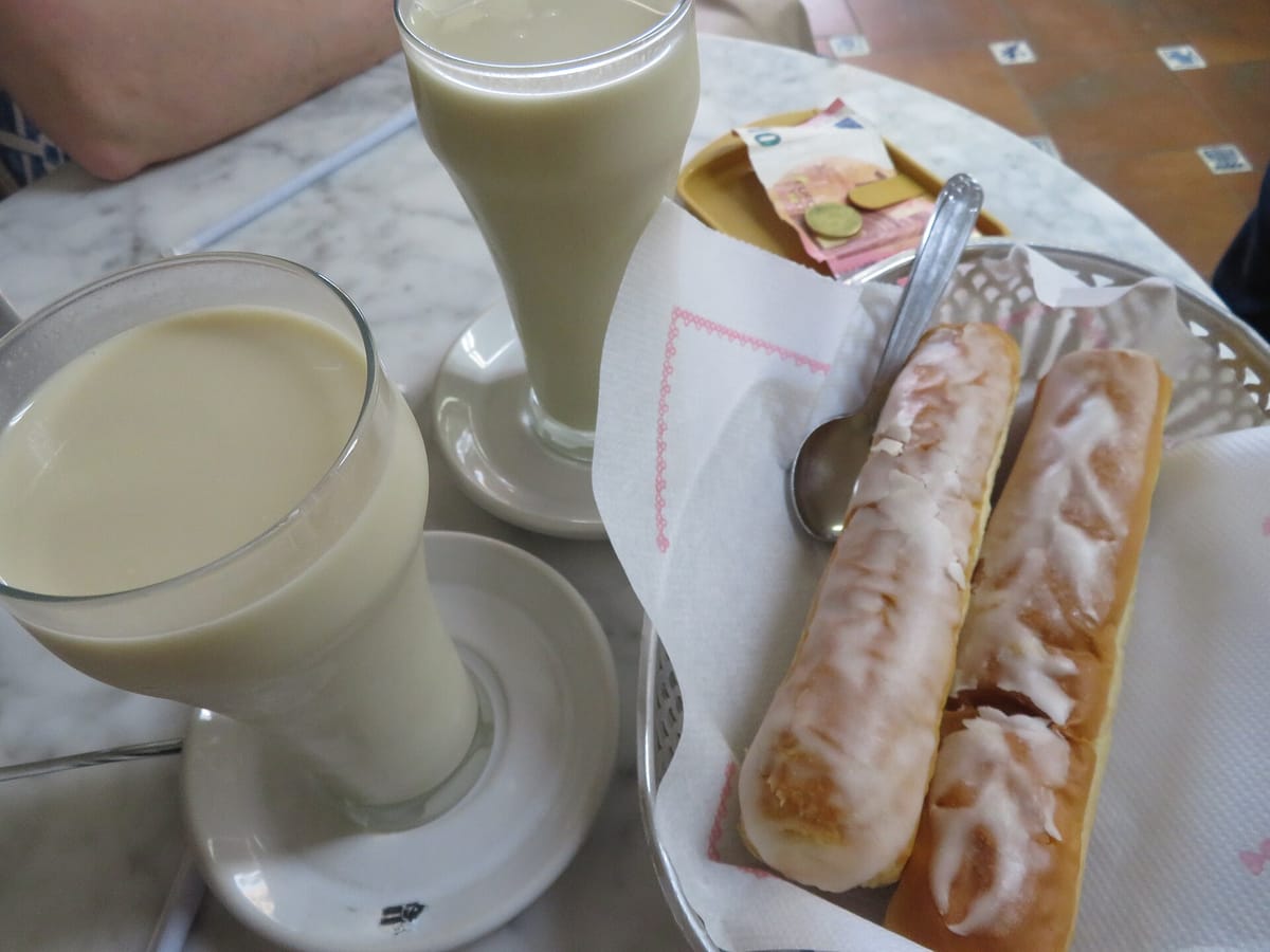 Two tall glasses of pale horchata on a marble cafe table in Valencia, beside a basket of two sugar-glazed fartons and a saucer with the bill and some coins